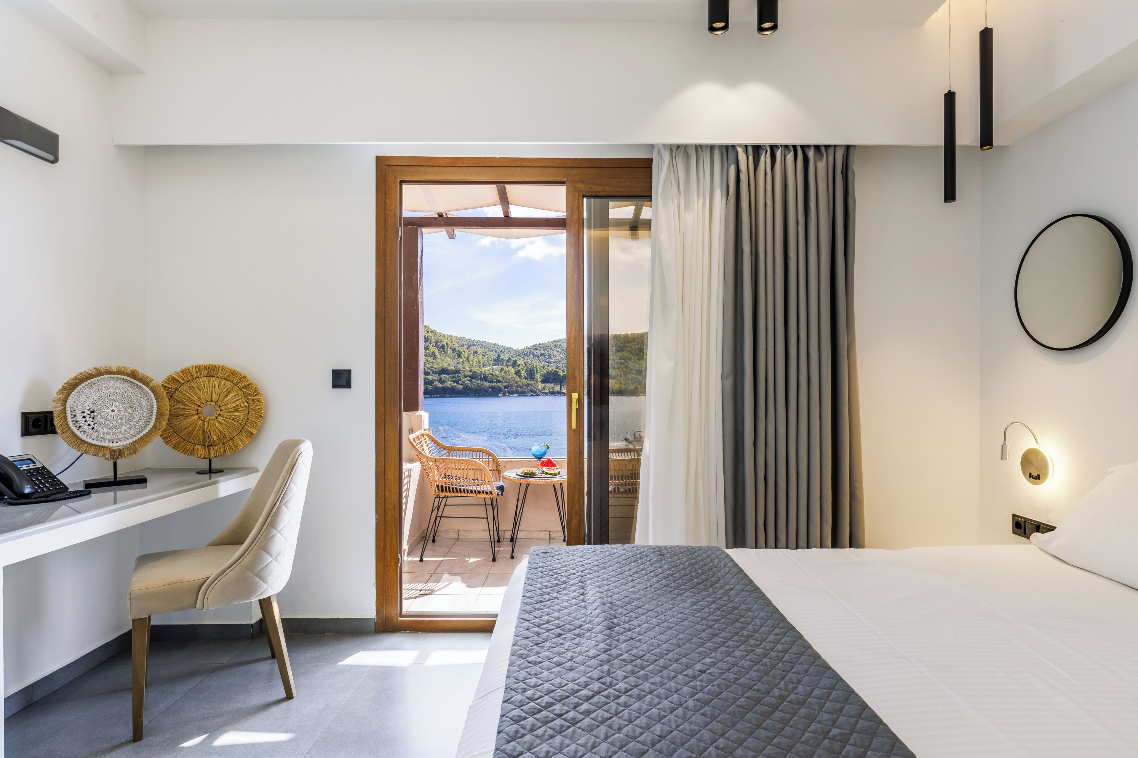 Una camera d'albergo con un letto, una scrivania con il telefono, due lampade a parete, vista sul mare e un balcone con tavolo e sedie.
