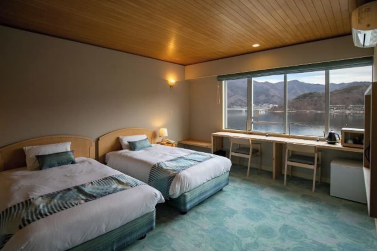 A hotel room with two beds, a desk, a chair, and a view of mountains through a large window.