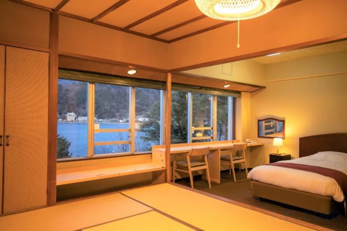 A traditional Japanese-style room with tatami flooring, a bed, a desk, and a view of a lake.