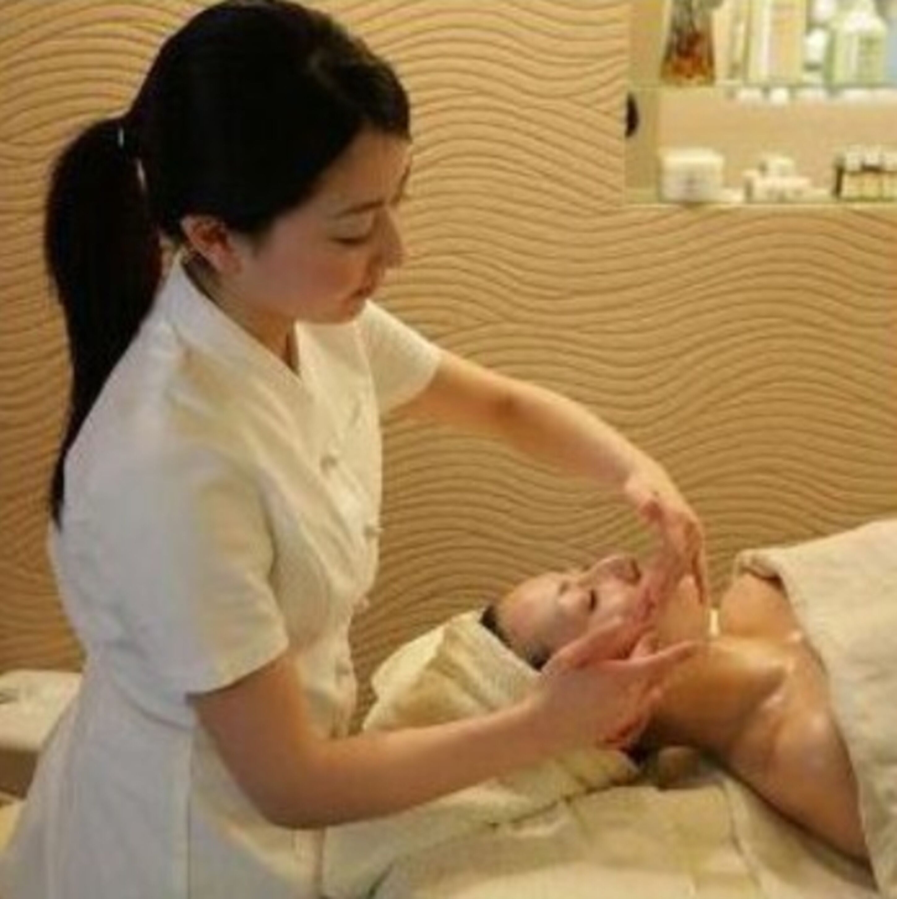 A person receiving a facial treatment in a room with a textured wall and a bed.