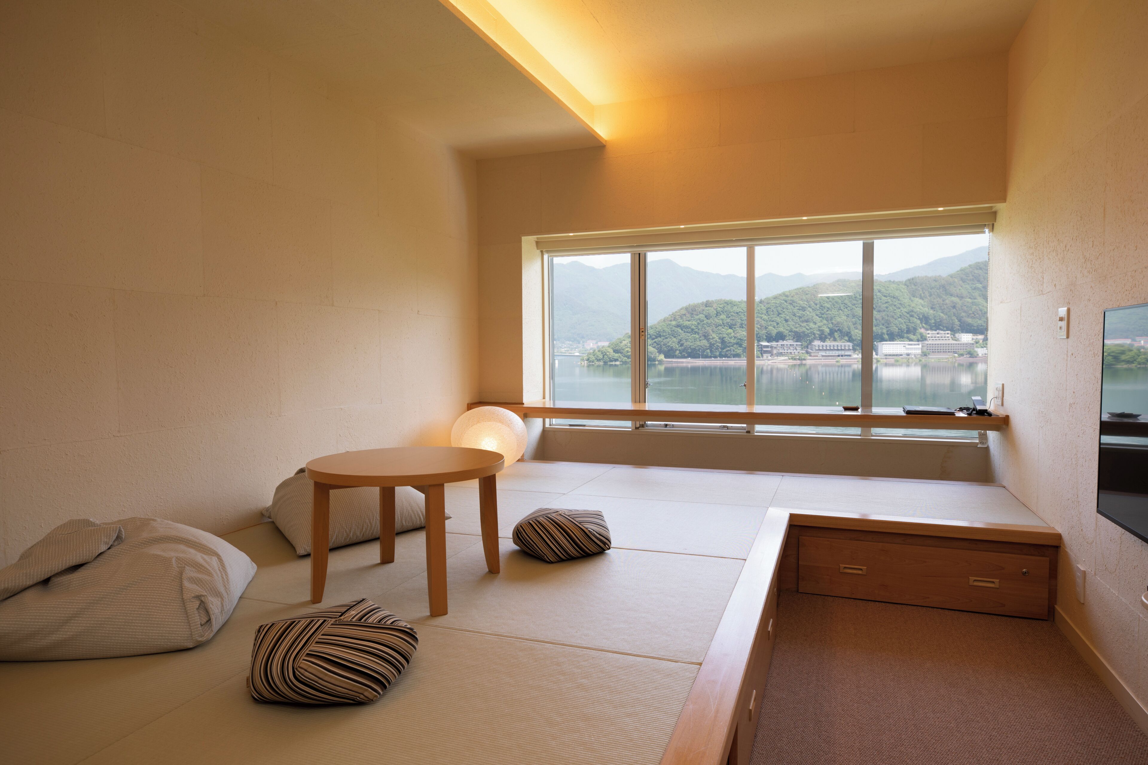 Una habitación con suelo de tatami, una mesa de madera baja, cojines, un televisor y un ventanal grande con vistas a un cuerpo de agua y colinas.