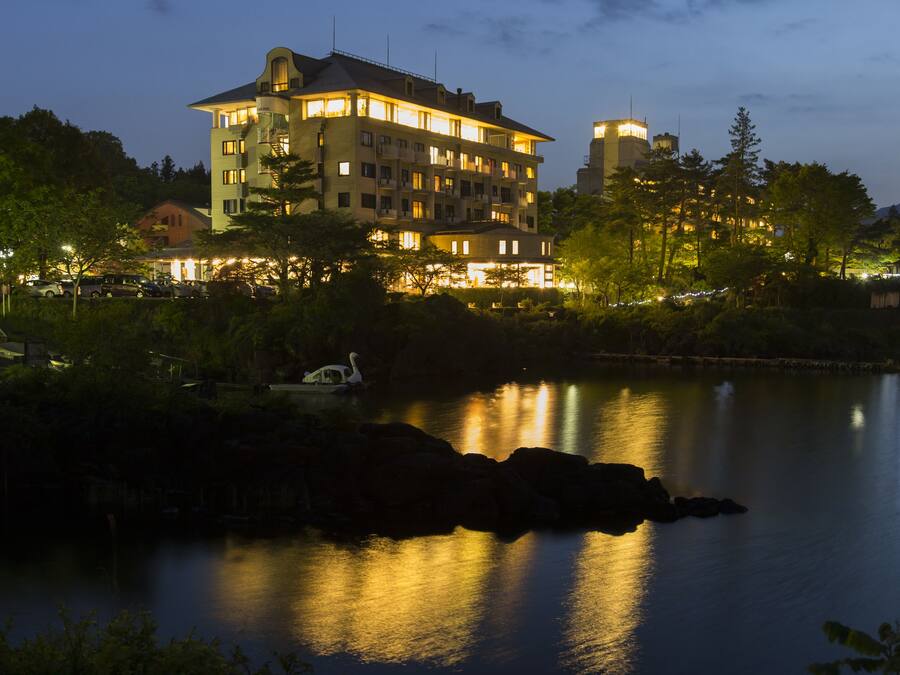 A multi-story hotel with balconies, a lake in the foreground, and a mountain in the background.