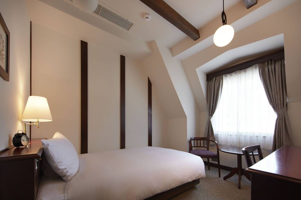 A hotel room with a bed, a nightstand with a lamp, a chair, a table, and a window with curtains.