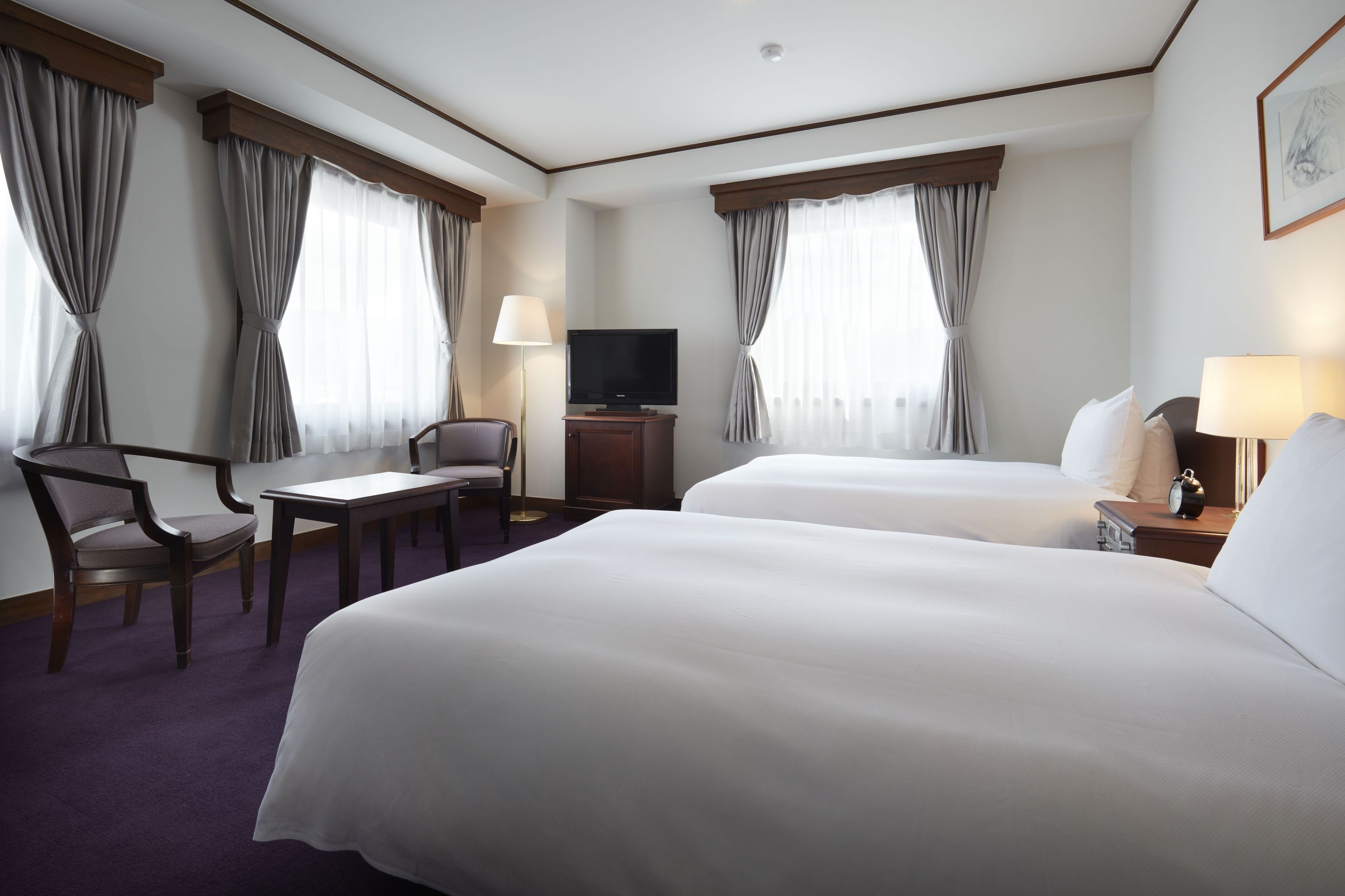 A hotel room with two beds, a desk, a chair, a television, and a lamp.