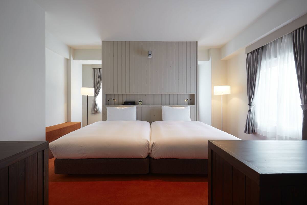 A hotel room with a double bed, a bedside table, a nightstand, a lamp, a window with curtains, and a wall-mounted control panel.