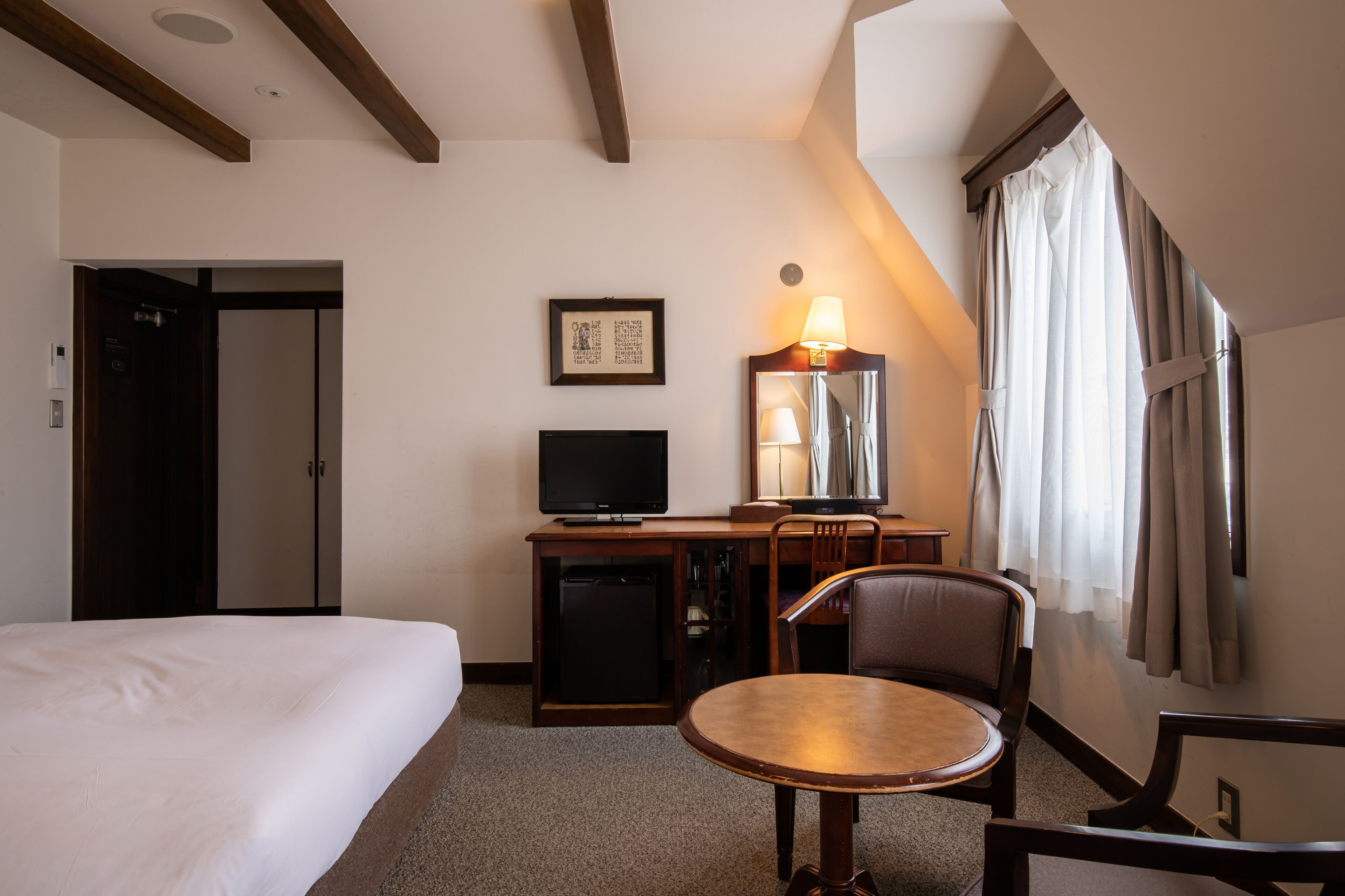 A hotel room with a bed, a desk with a television, a chair, a lamp, and a window with curtains.
