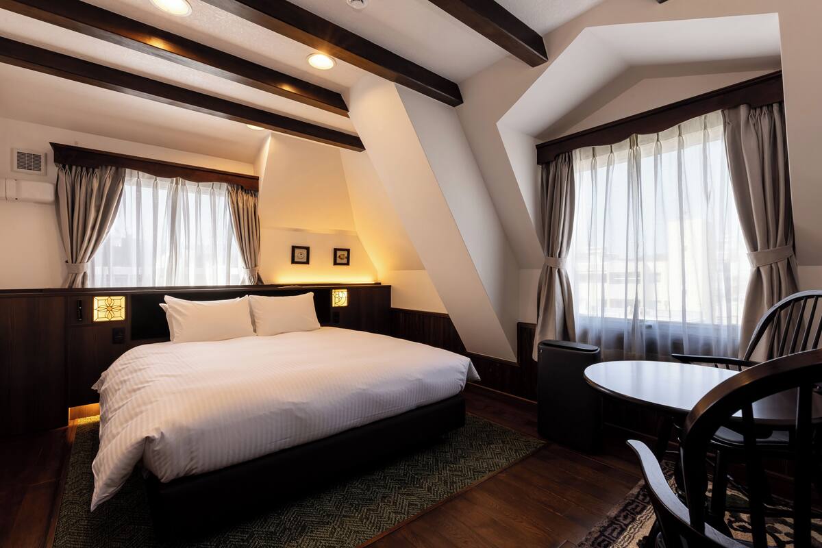 A bedroom with a bed, bedside tables, a chair, a small table, and a window with curtains.