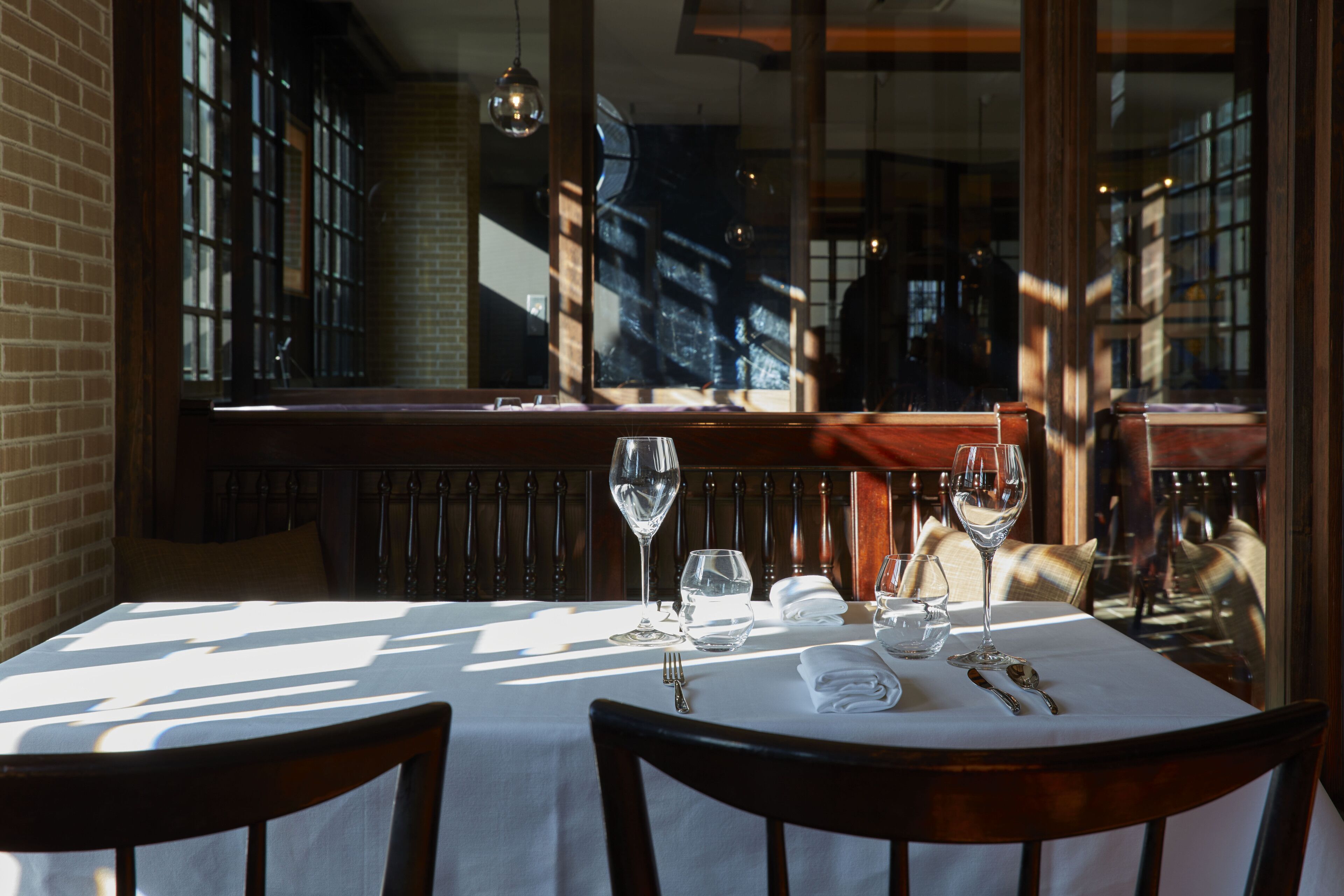 A dining table set with wine glasses, water glasses, and cutlery, with a view of a brick wall and large windows in the background.