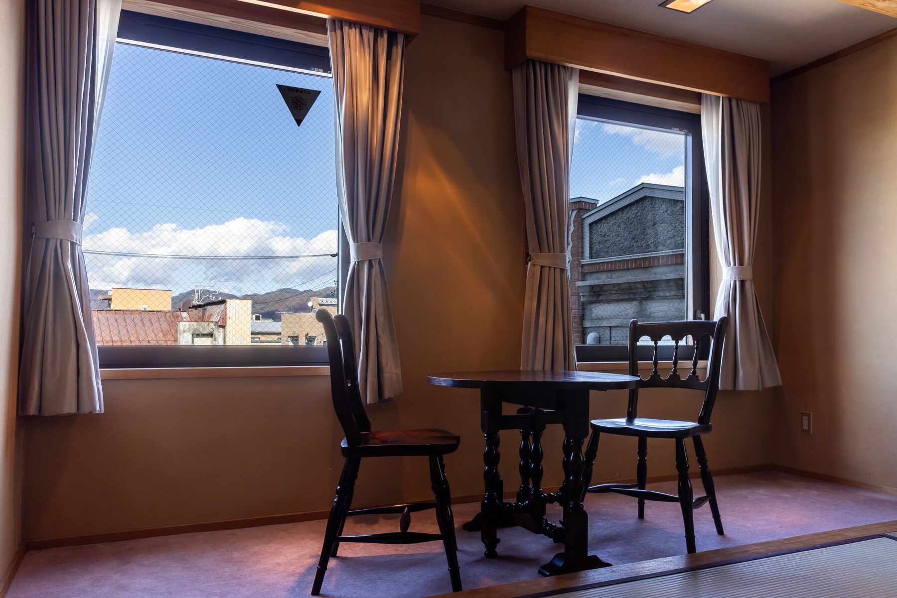 Una habitación con una mesa de comedor y sillas, ventanas grandes con cortinas y vistas a edificios y montañas.