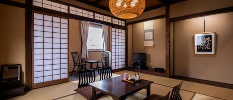 A traditional Japanese room with tatami flooring, a low table with cushions, a television, and a hanging lamp.