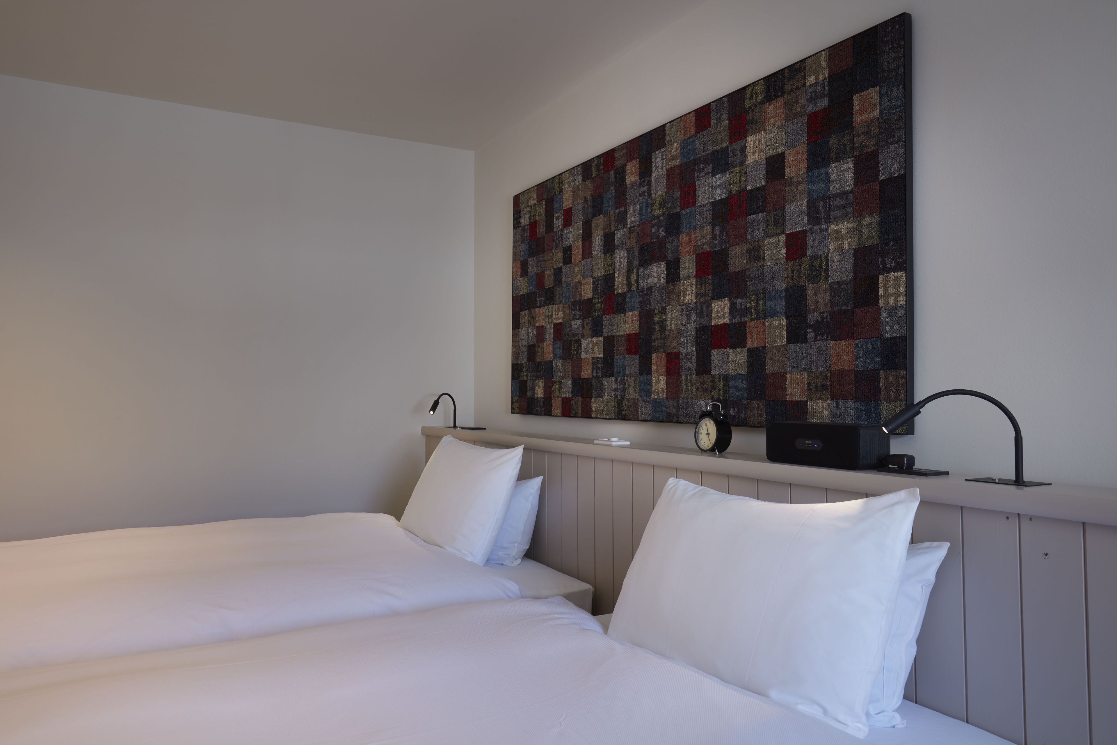 A hotel room with two beds, a wall-mounted artwork, a bedside table with a lamp, and a clock.
