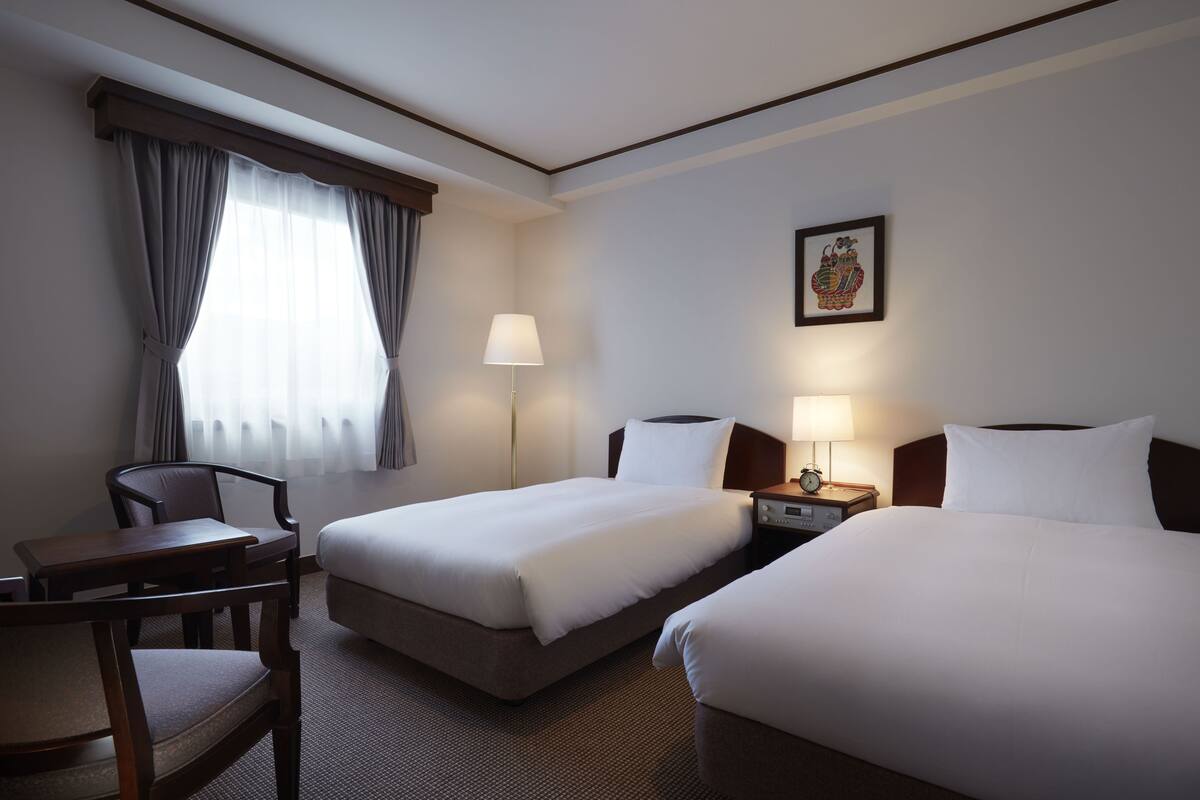 A hotel room with two beds, a desk, a chair, a nightstand with a lamp, and a window with curtains.