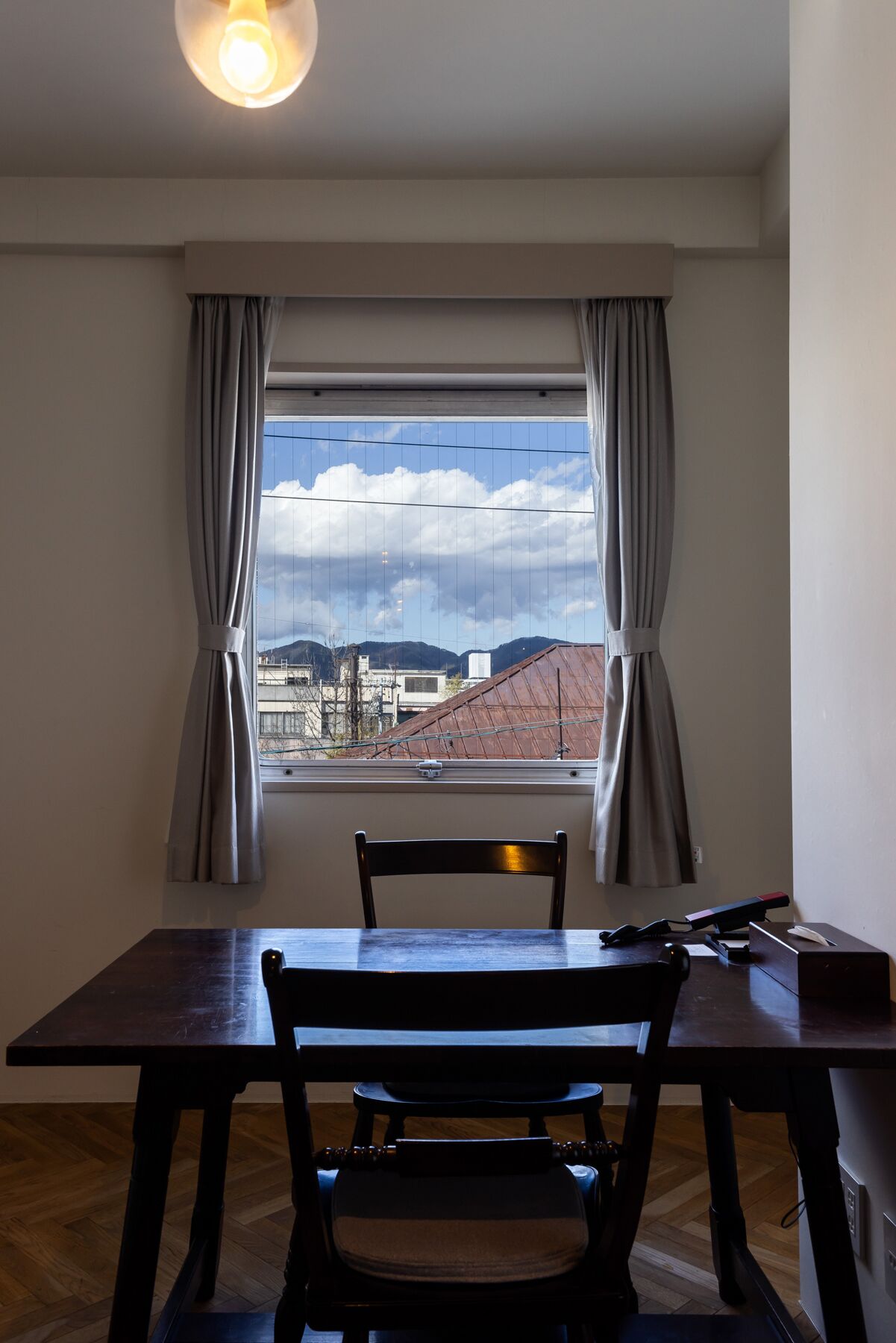 A room with a dining table and chairs, a window with curtains, and a view of buildings and mountains.