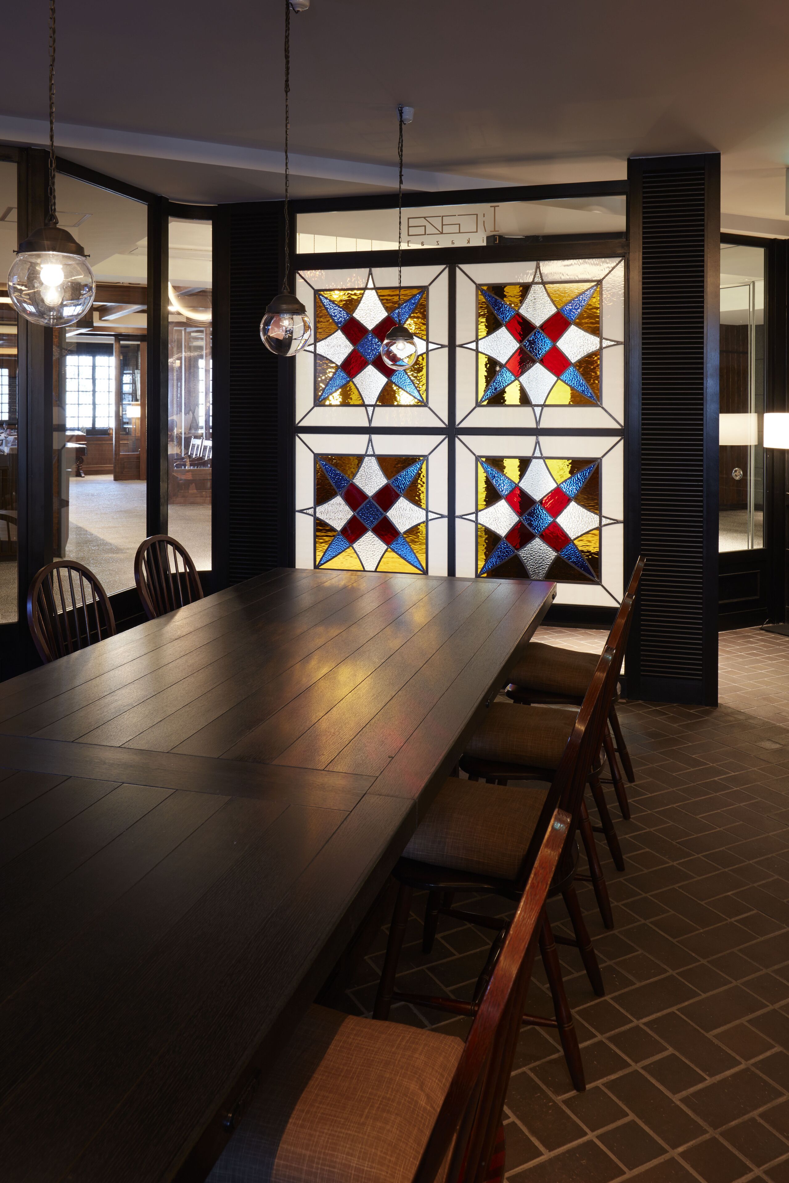 A dining area with a long wooden table, several chairs, and a stained glass window with geometric patterns.