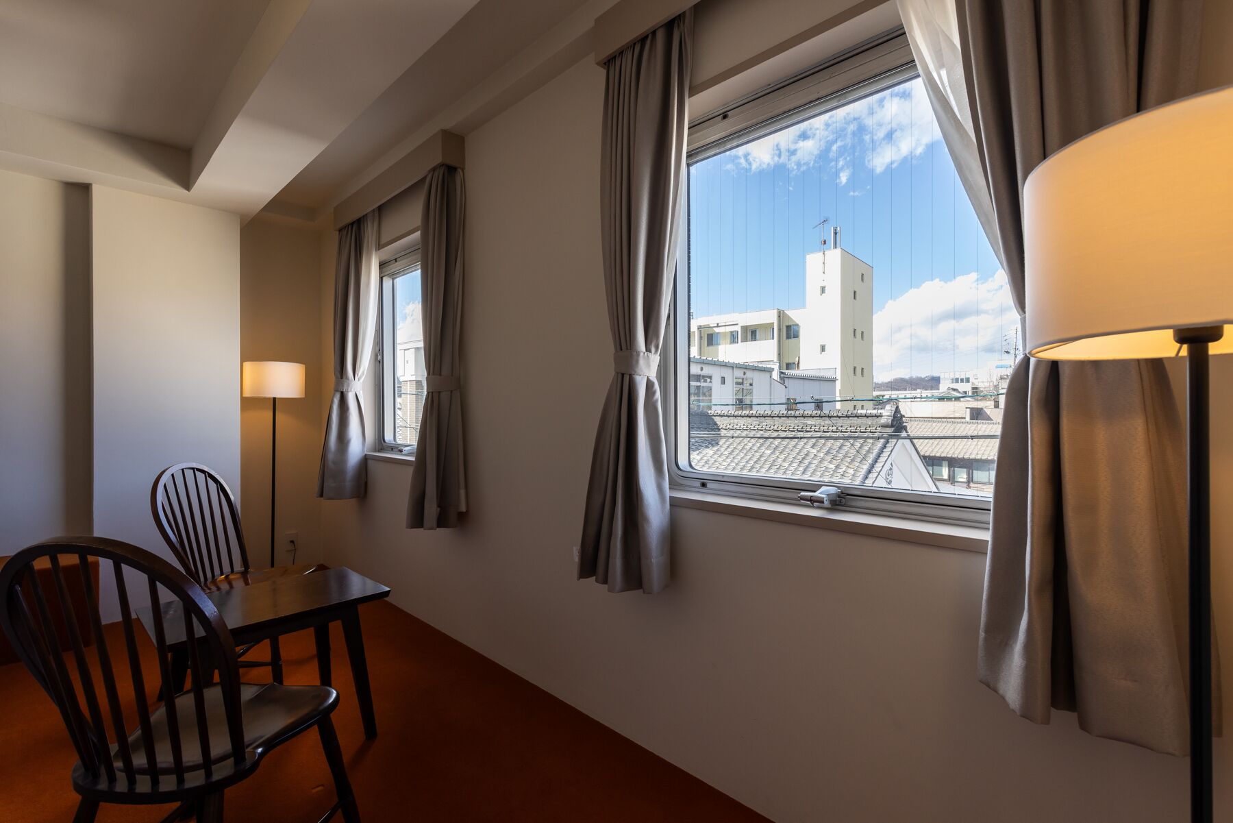 Habitación de hotel con una mesa de comedor, dos sillas, una lámpara y un ventanal con cortinas.