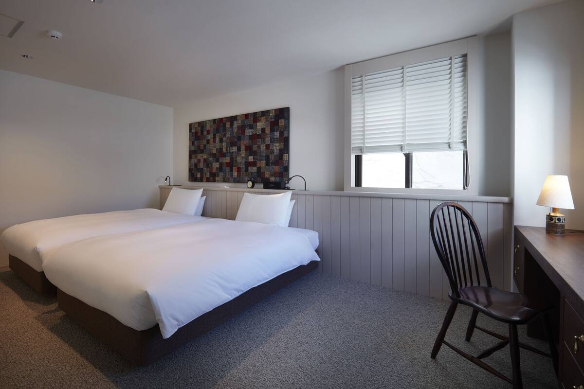 A bedroom with a large bed, a desk, a chair, a window with blinds, and a wall-mounted clock.