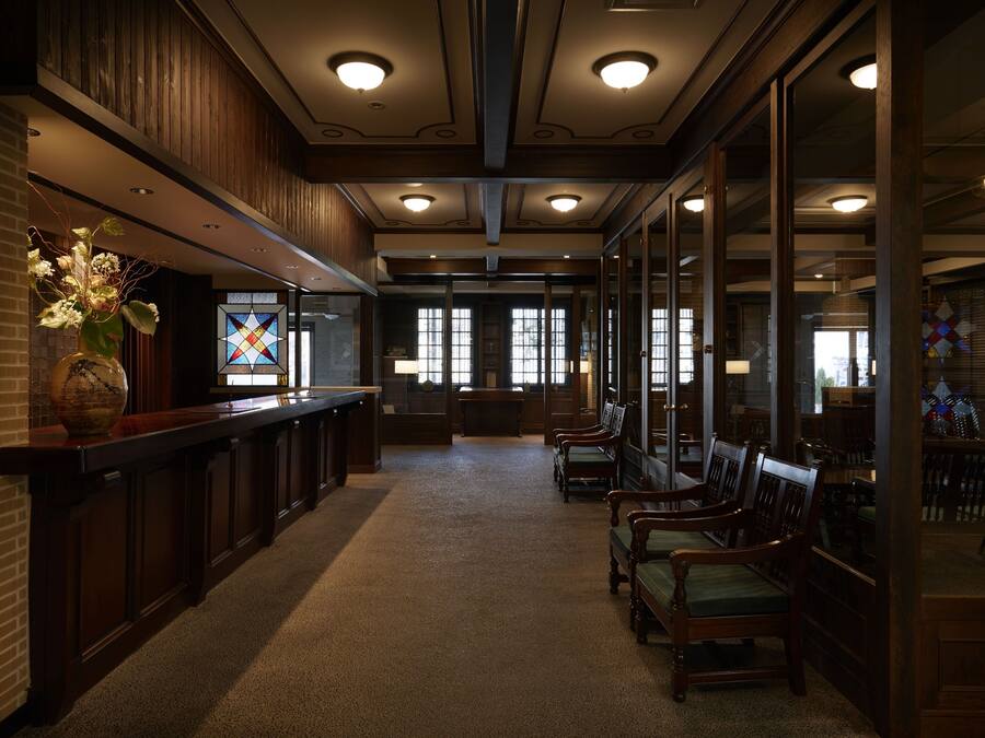 A reception area with a wooden reception desk, a vase with flowers, and a stained glass window. There are several wooden chairs and tables arranged for seating.