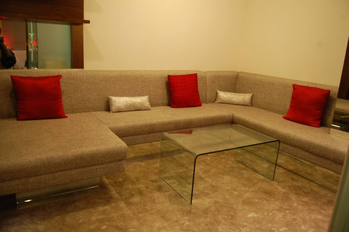 Ein modernes Wohnzimmer mit einem beigefarbenen Ecksofa, zwei roten und zwei weißen Kissen sowie einem Glastisch.