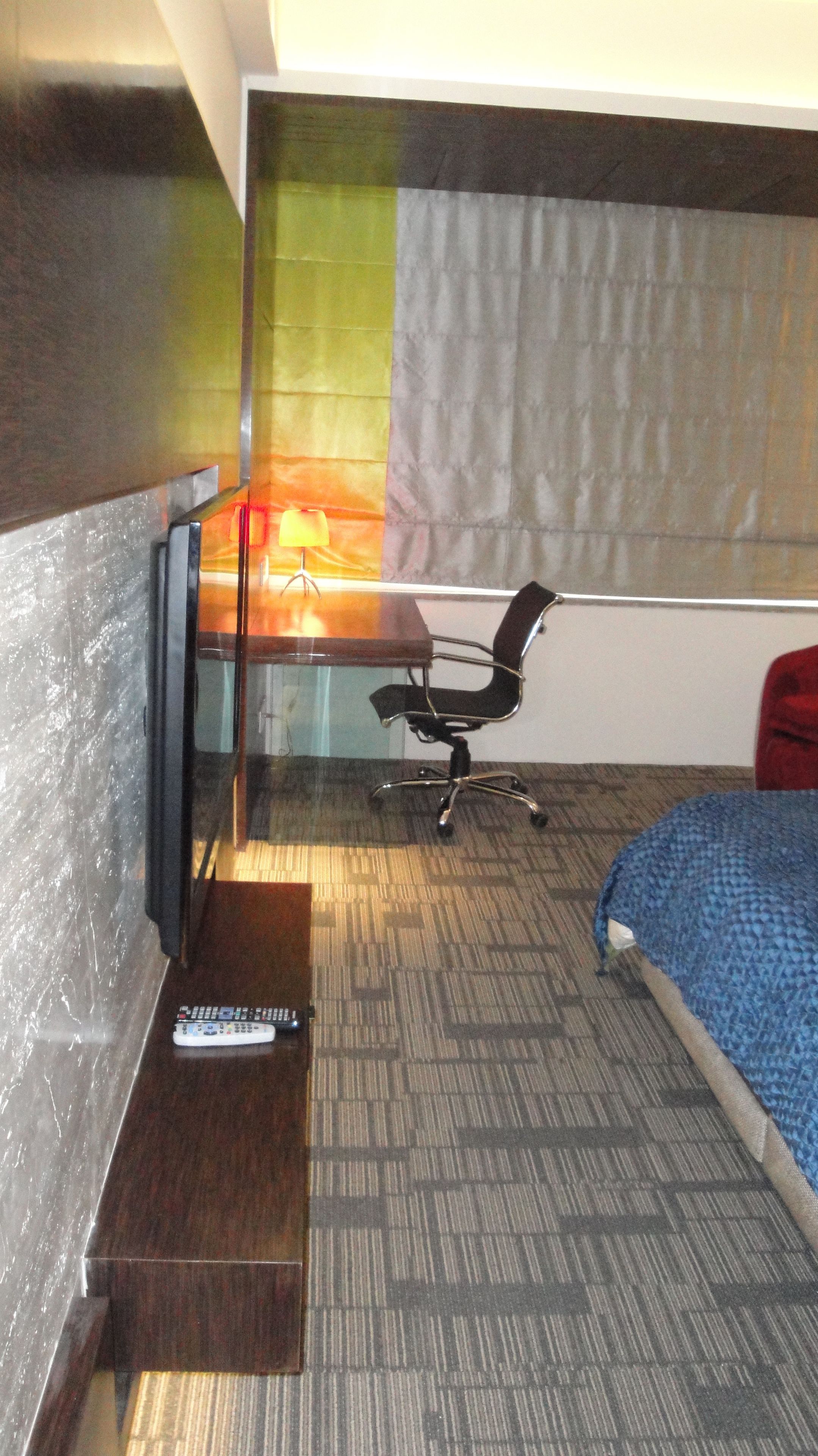 Habitación de hotel con cama, escritorio con silla, control remoto y ventana con cortinas.