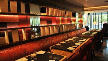 Restaurante com mesas compridas, bancos estofados vermelhos e parede estampada com painéis iluminados.