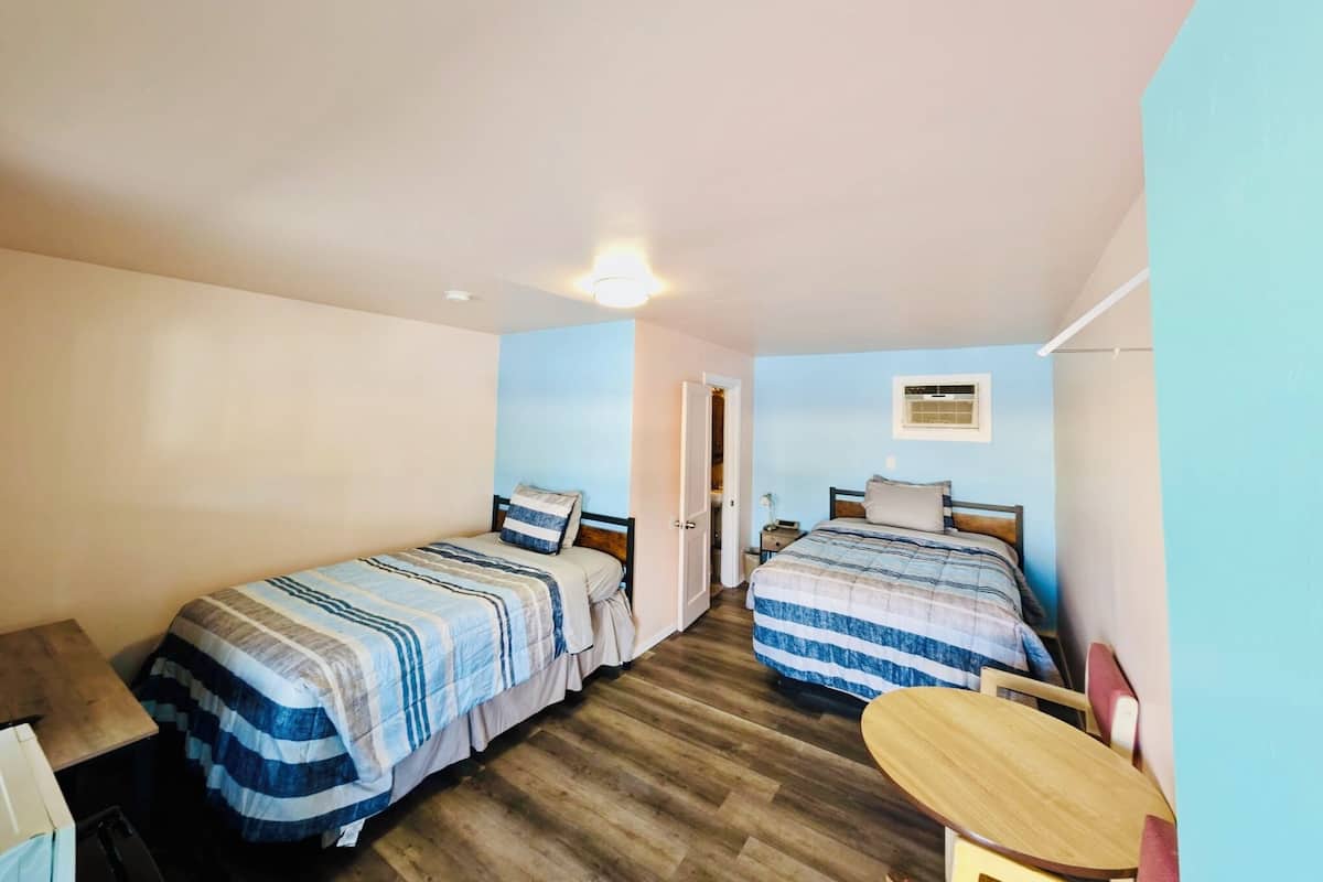 A small, well-lit room with two beds, a wooden floor, a small table, and a door leading to another room.