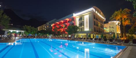 A swimming pool with lounge chairs and umbrellas, a building with balconies, and a mountain in the background.