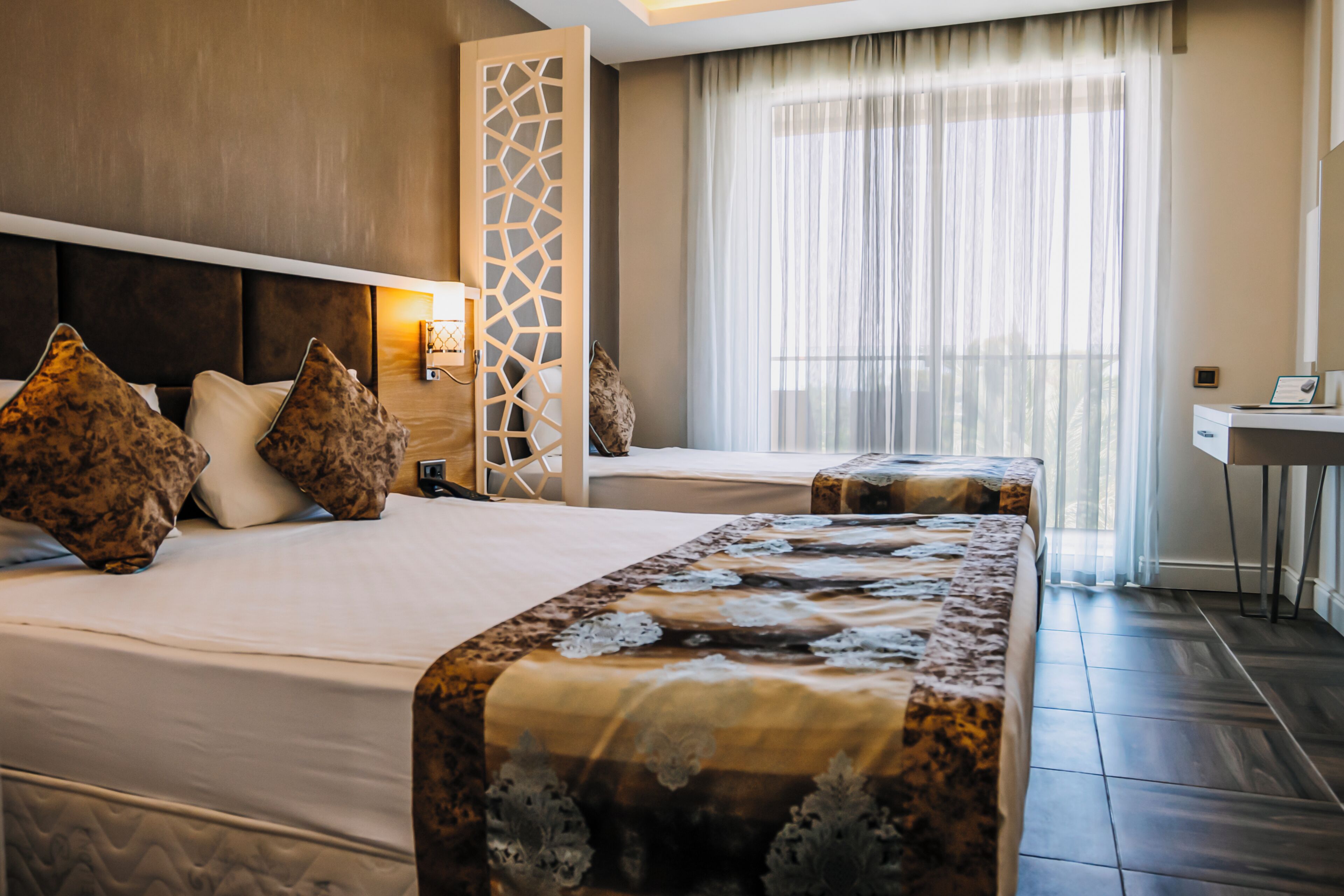 Une chambre d’hôtel avec deux lits, un bureau, une chaise, un panneau décoratif au mur et une grande fenêtre avec des rideaux.