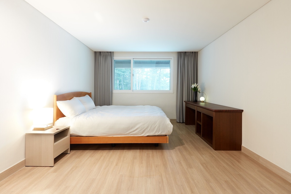 A modern bedroom with a wooden bed, a bedside table, a nightstand, a desk, and a window with curtains.