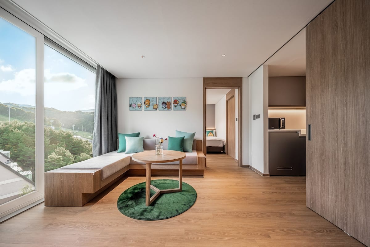 Ein modernes Wohnzimmer mit großem Fenster, einer Couch mit türkisfarbenen Kissen, einem kleinen runden Tisch und einem grünen Teppich.