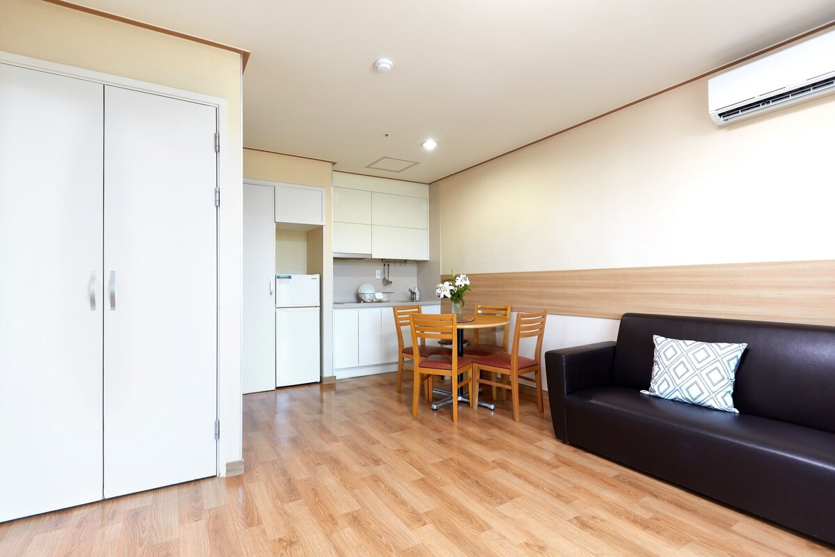 A modern kitchen with white cabinets, a dining table with chairs, and a black leather sofa with a patterned pillow.