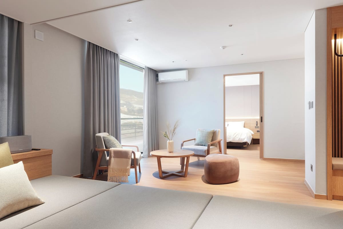 Ein modernes Hotelzimmer mit einem großen Bett, einer Couch, einem runden Couchtisch und einer Sitzecke mit Stühlen. Es gibt ein großes Fenster mit Vorhängen, eine Klimaanlage und einen Blick ins Freie.
