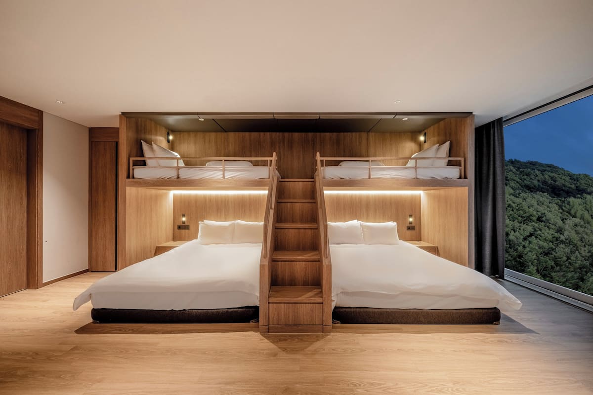 Ein modernes Schlafzimmer mit zwei Betten, einer Holzleiter und einem großen Fenster mit Blick auf eine bewaldete Landschaft.