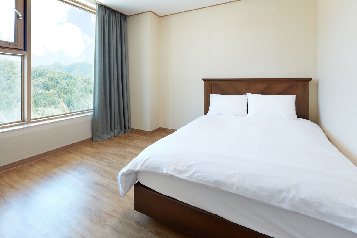 A neatly made bed in a room with a large window, wooden flooring, and a view of a forested area.