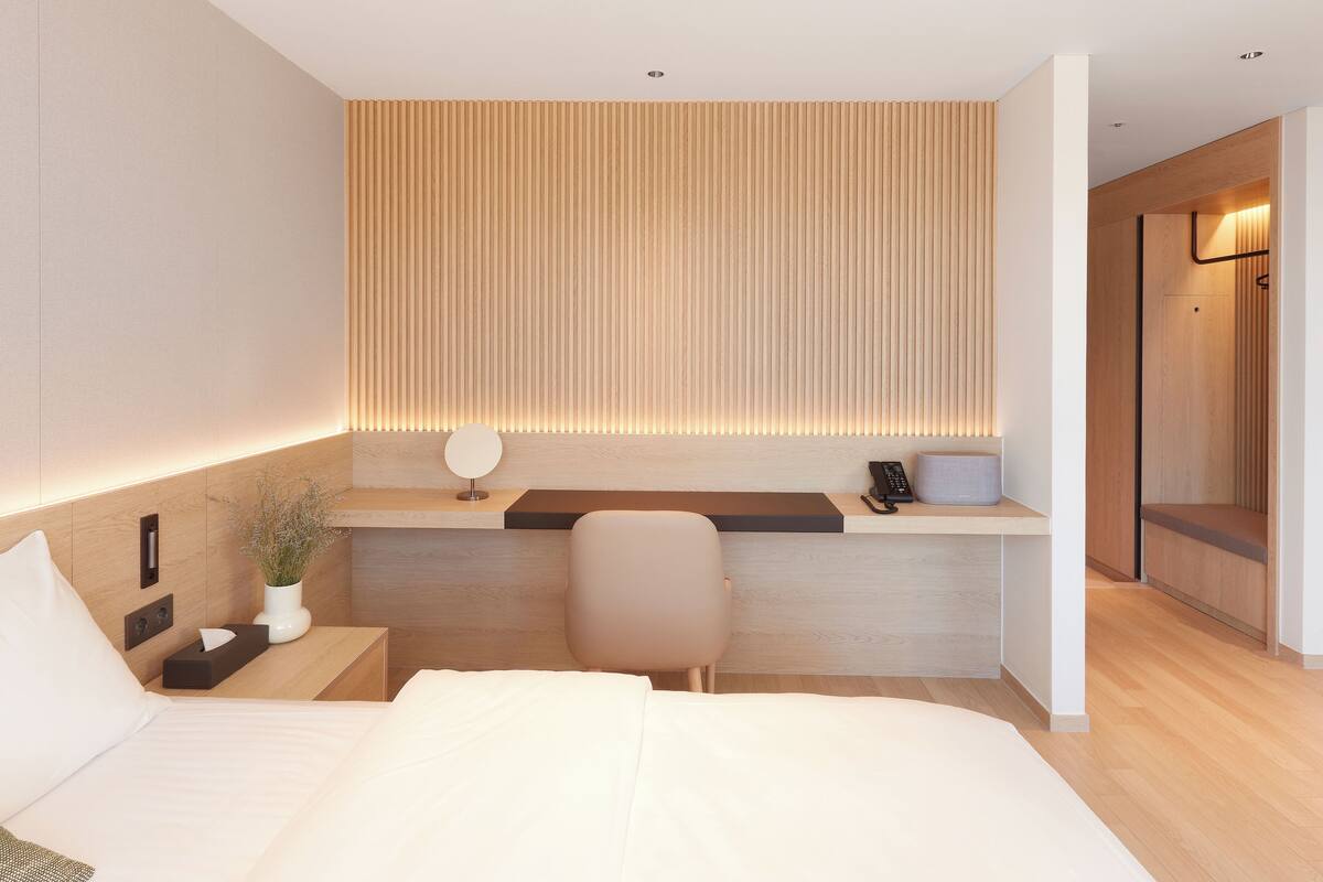 Ein modernes Hotelzimmer mit Bett, Nachttisch, Schreibtisch mit Stuhl, Wandleuchte und einer Wandverkleidung aus Holz.