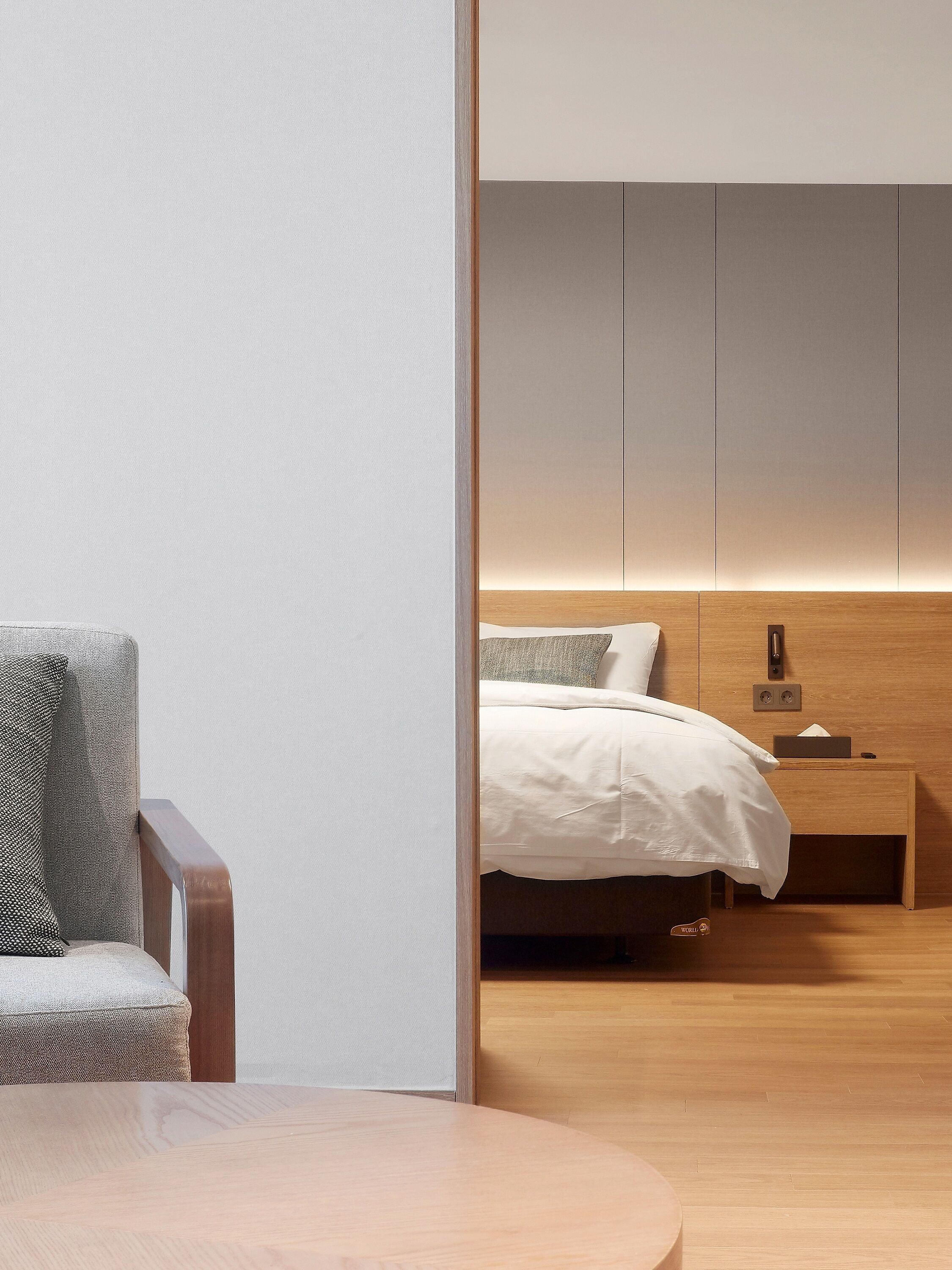 A modern hotel room with a wooden floor, a bed with white bedding, a nightstand with a lamp, and a chair with a patterned cushion.