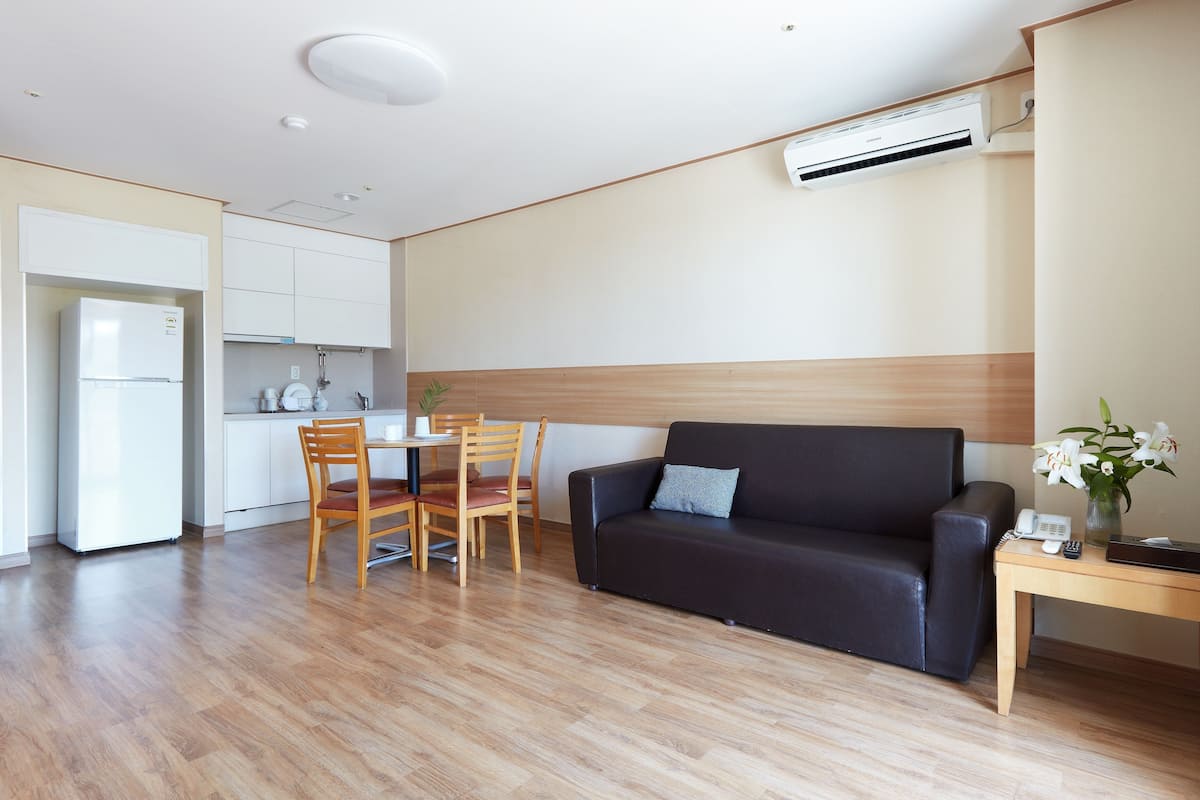 Ein modernes Wohnzimmer mit einem dunklen Sofa, Holzstühlen, einem kleinen Tisch mit Blumen und einer Küchenzeile mit weißen Schränken und einem Kühlschrank.