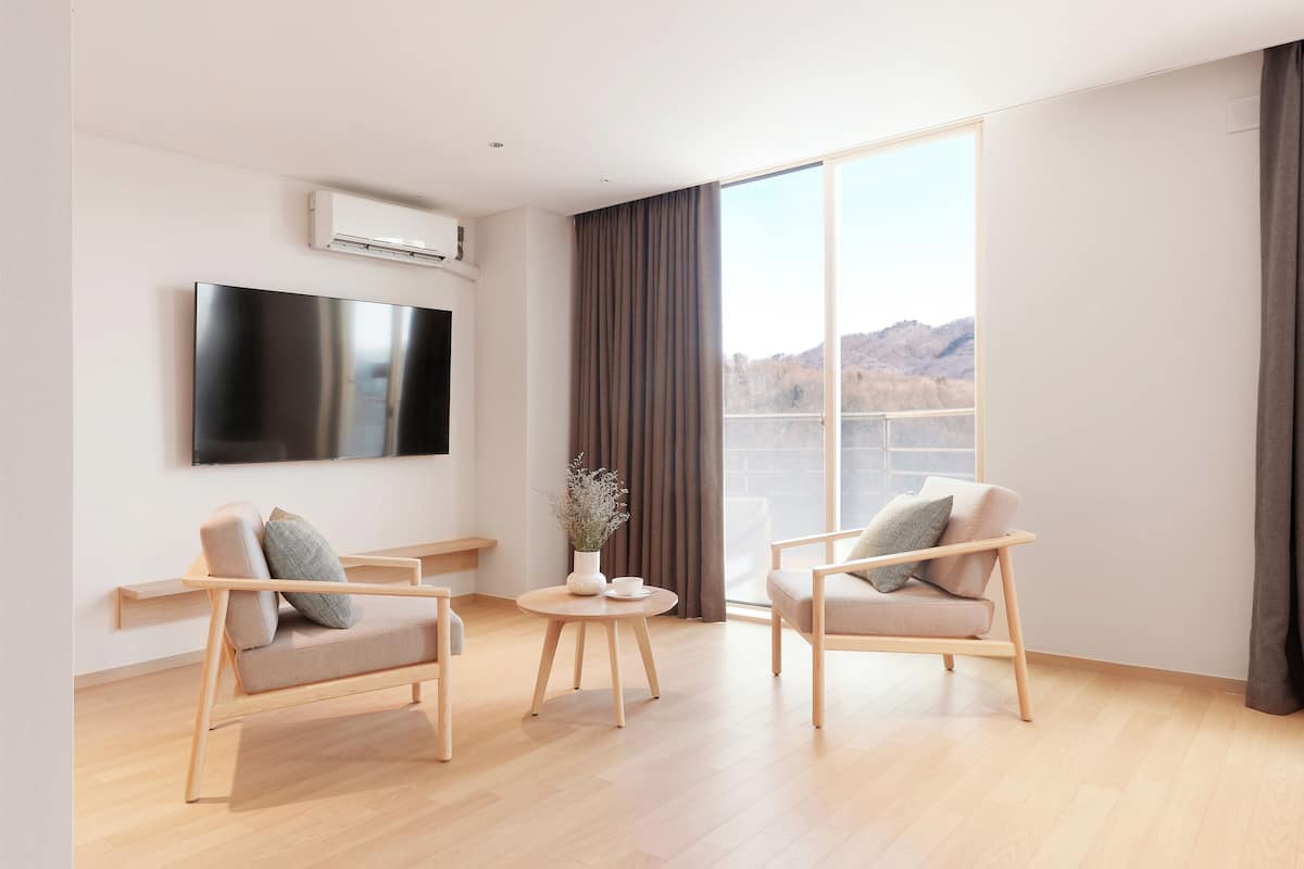 Ein modernes Wohnzimmer mit einem Flachbildfernseher, zwei Sesseln, einem kleinen runden Tisch mit einer Pflanze und einem großen Fenster mit Blick auf Hügel.