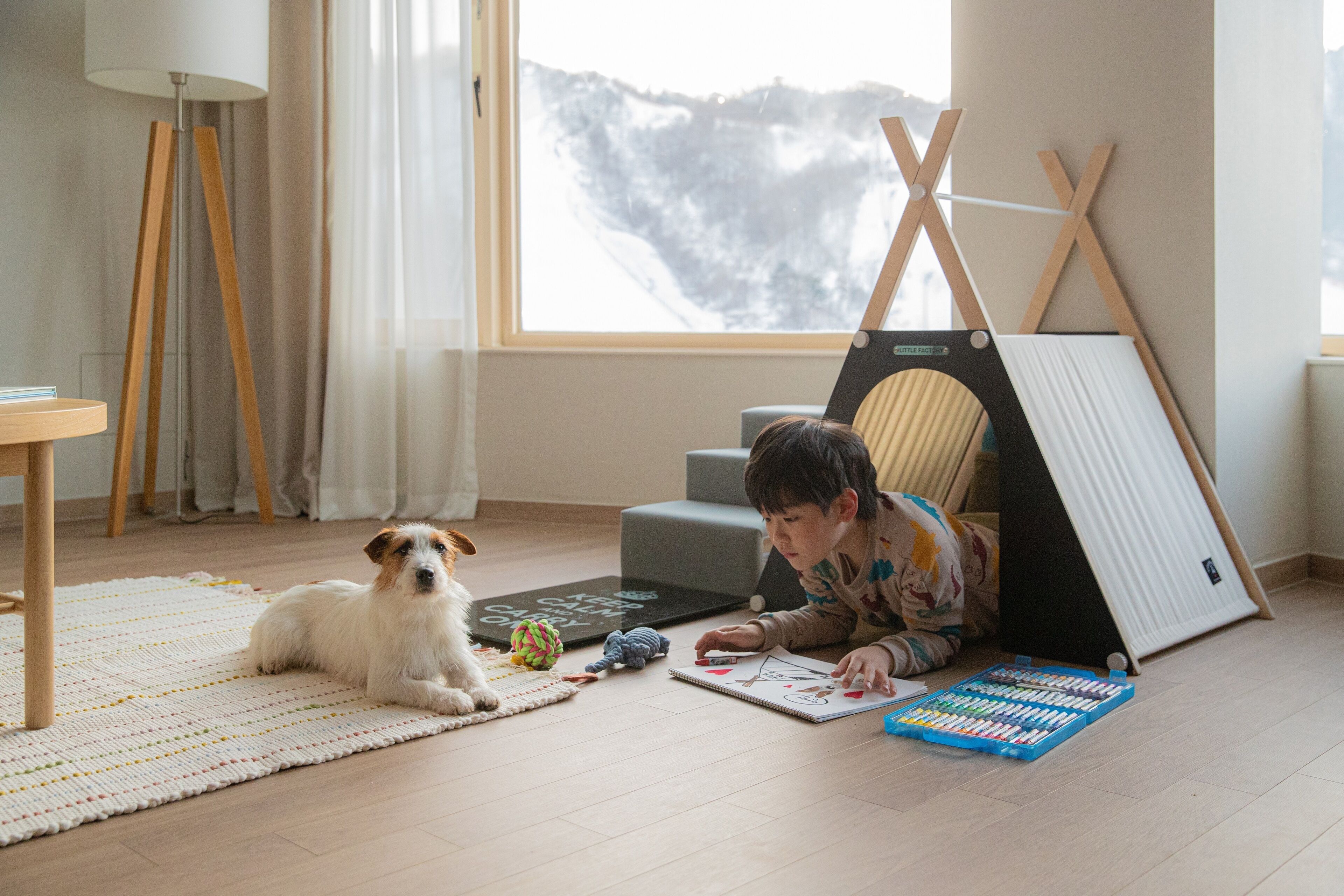 A child playing with a puzzle on a rug, a dog lying nearby, and a tent-like structure in a room with a large window.