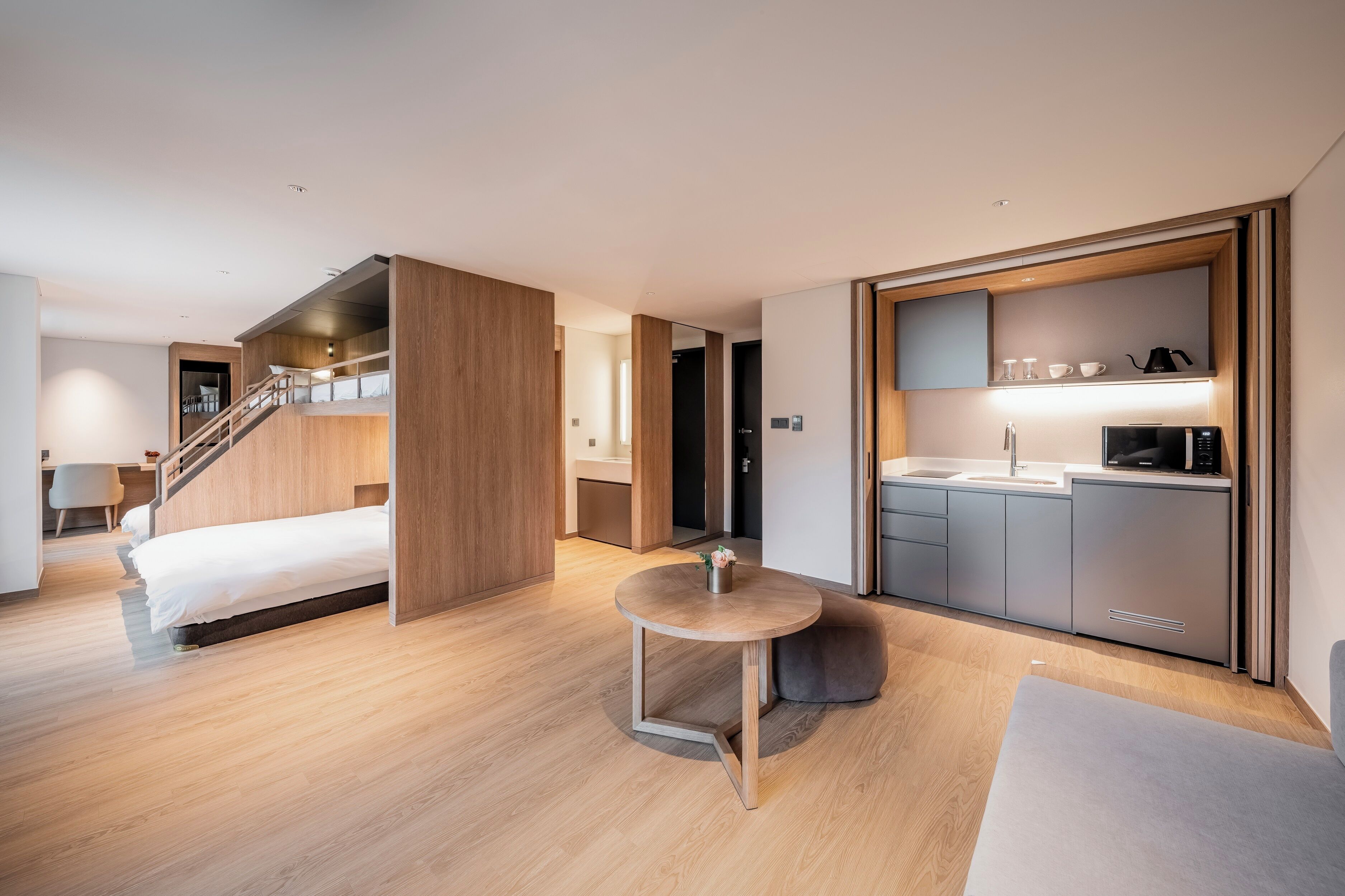 A modern hotel room with a bed, a small round table, a kitchenette, and a staircase leading to an upper level.