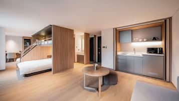 A modern hotel room with a bed, a small round table, a kitchenette, and a staircase leading to an upper level.