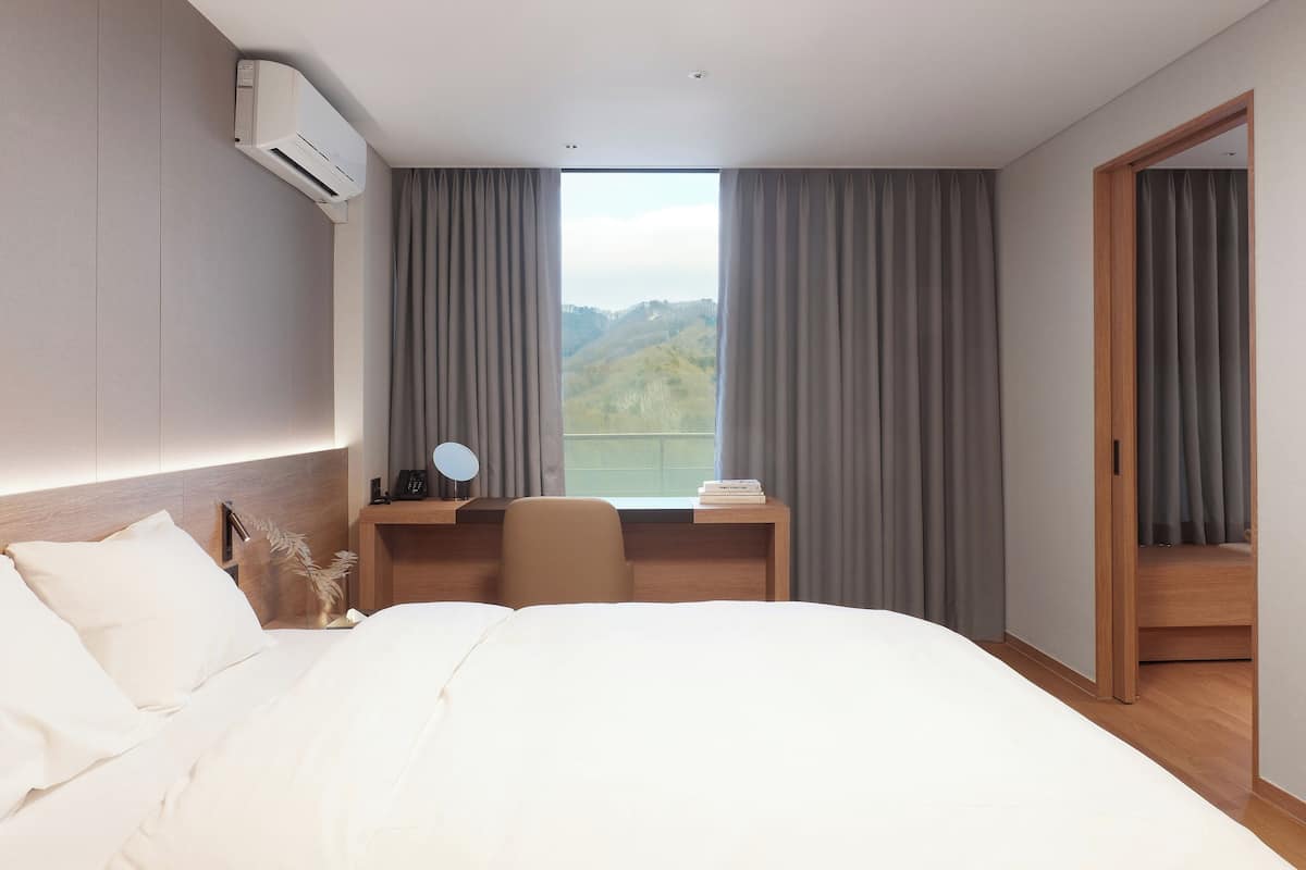 Ein Hotelzimmer mit einem großen Bett, einem Schreibtisch mit Stuhl, einem Fenster mit Vorhängen und einer Klimaanlage.