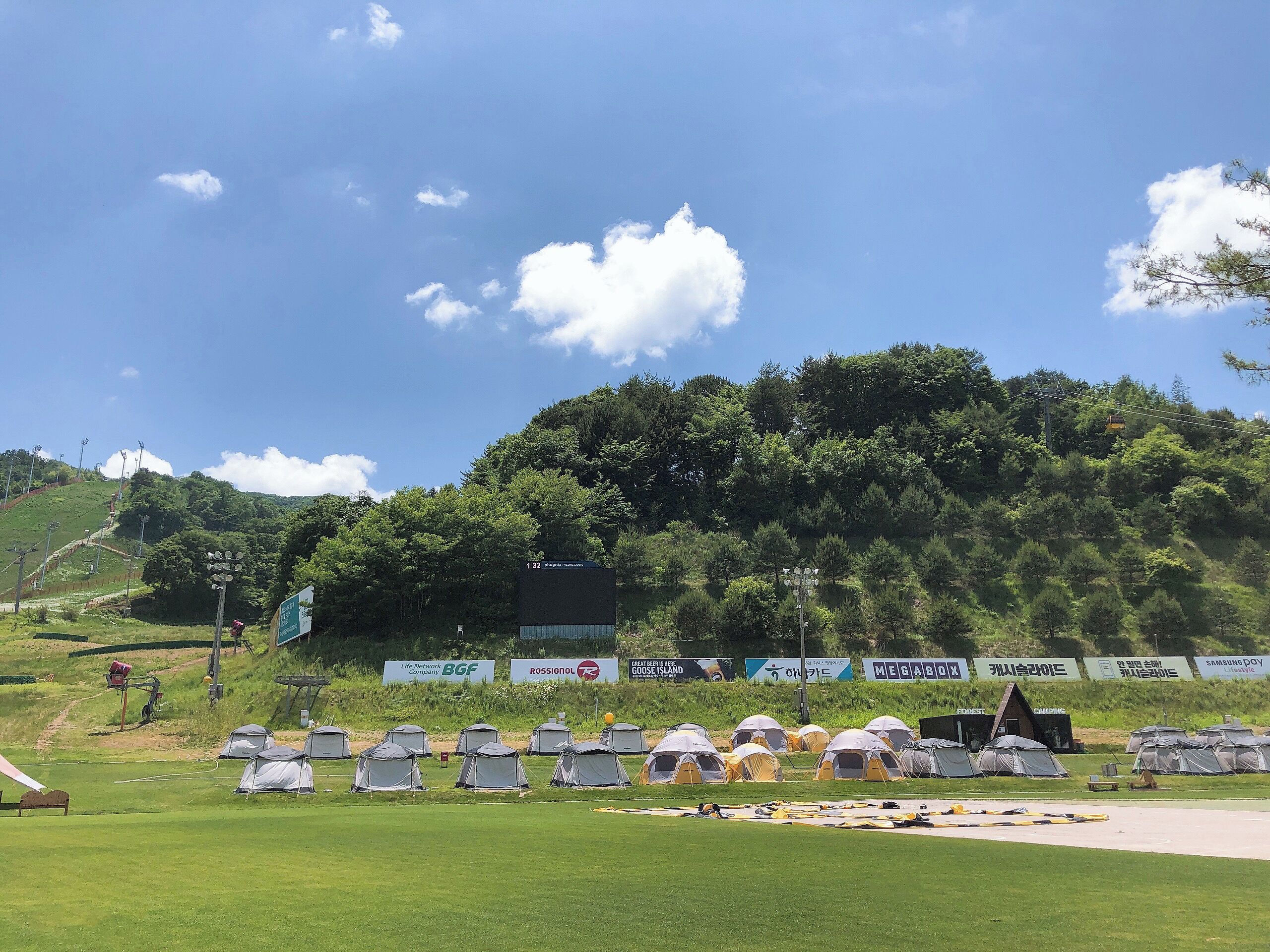 A campsite with tents, a grassy field, and a forested hill in the background.