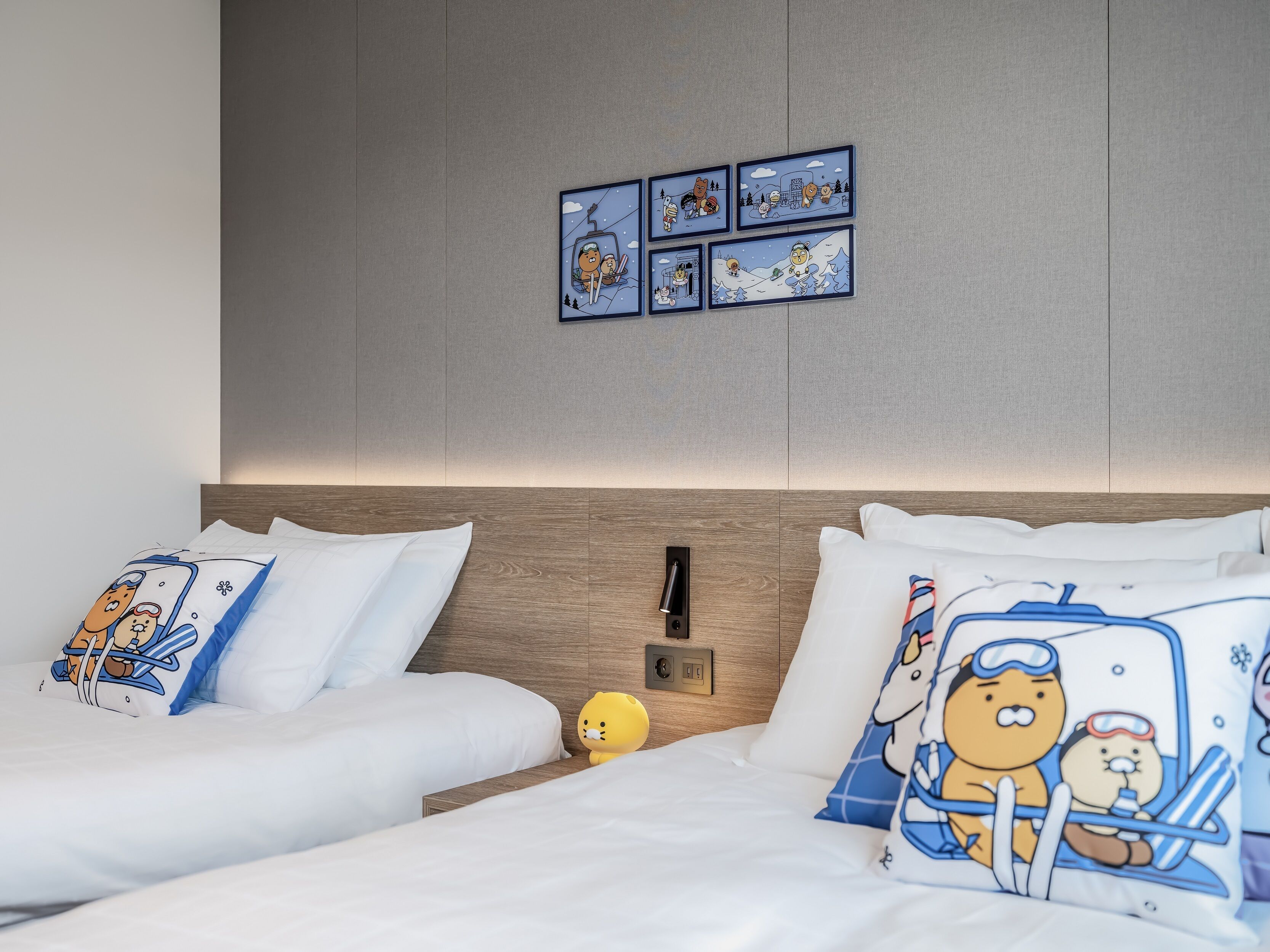 A hotel room with two beds, each with white bedding and cartoon-themed pillows. There is a wall-mounted light fixture and a small nightstand with a lamp.