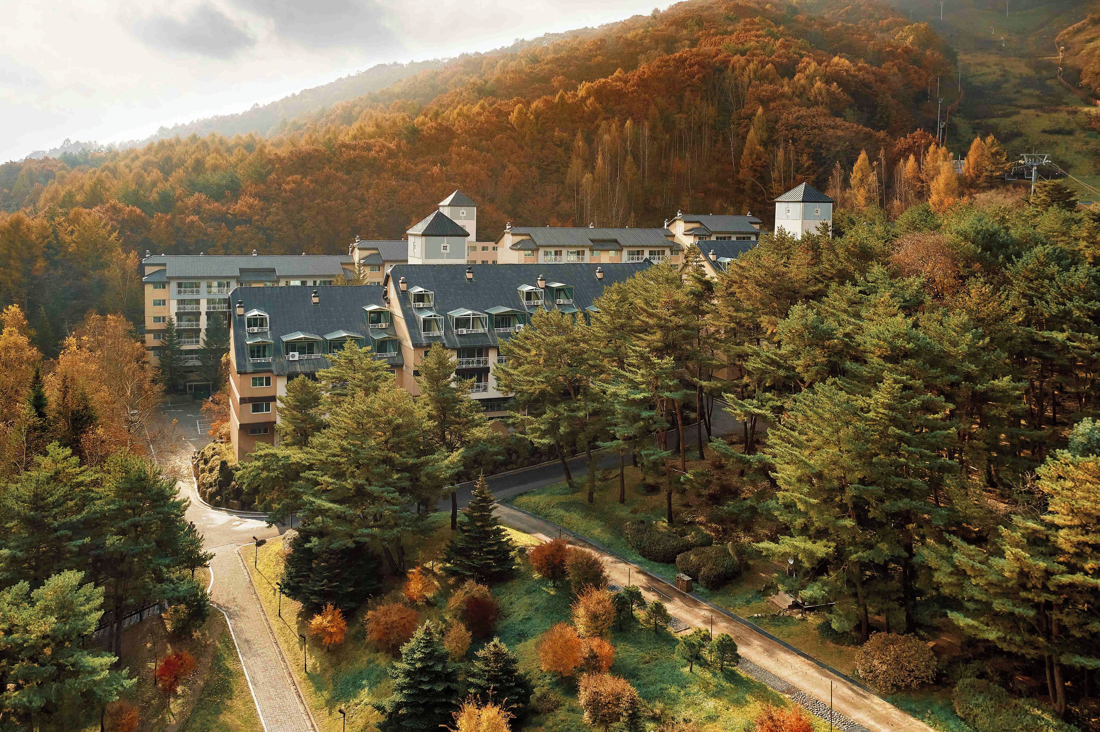 Un immeuble à plusieurs étages doté d’un parking et d’une route y menant, le tout entouré d’arbres et de collines.