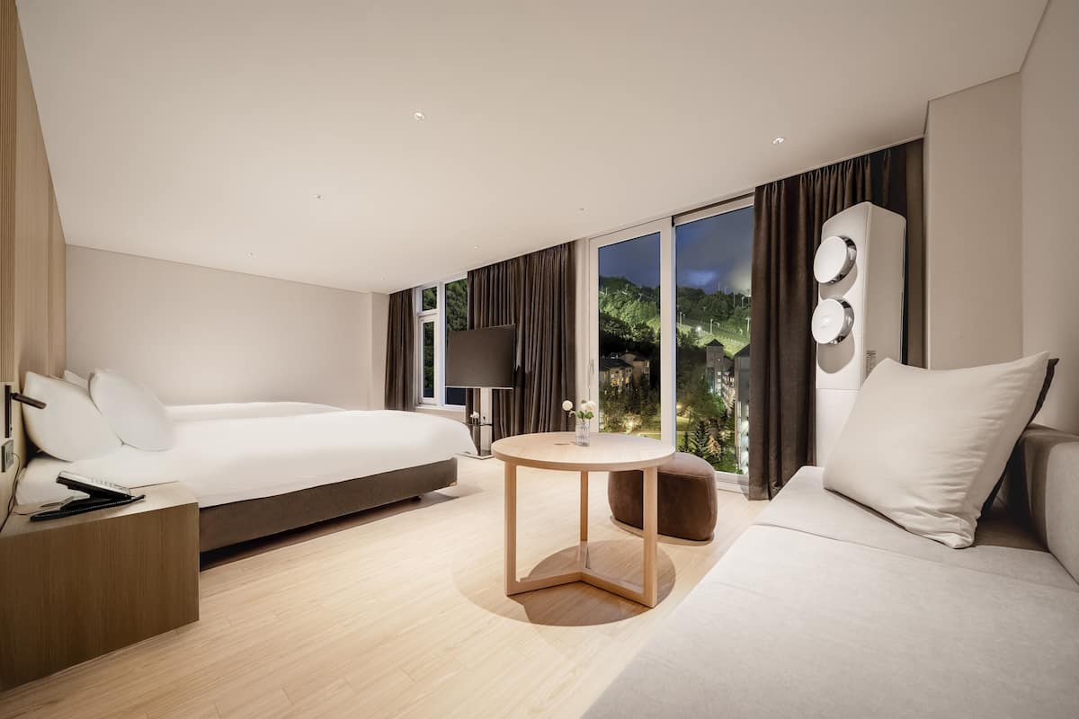 Ein modernes Hotelzimmer mit Bett, Sofa, einem kleinen runden Tisch und einem großen Fenster mit Blick ins Freie.