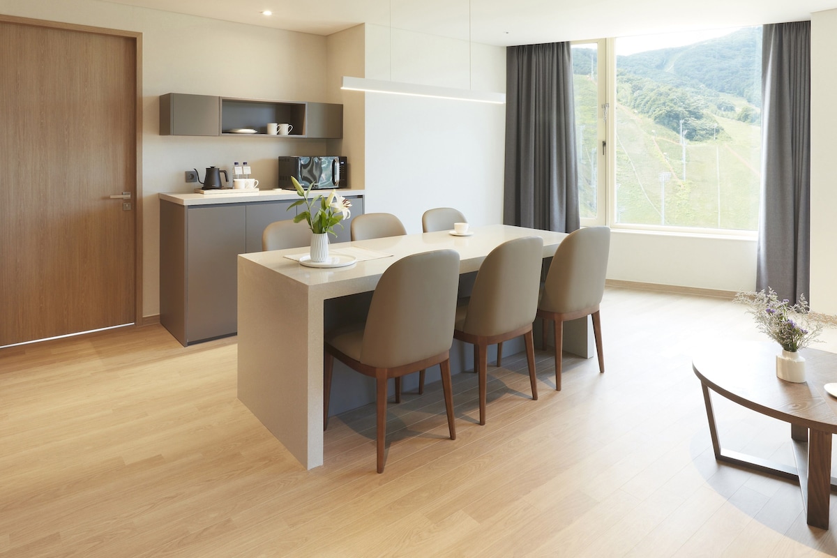 A modern kitchen with a microwave and cabinets, a dining area with a table and chairs, and a view of a green landscape through large windows.