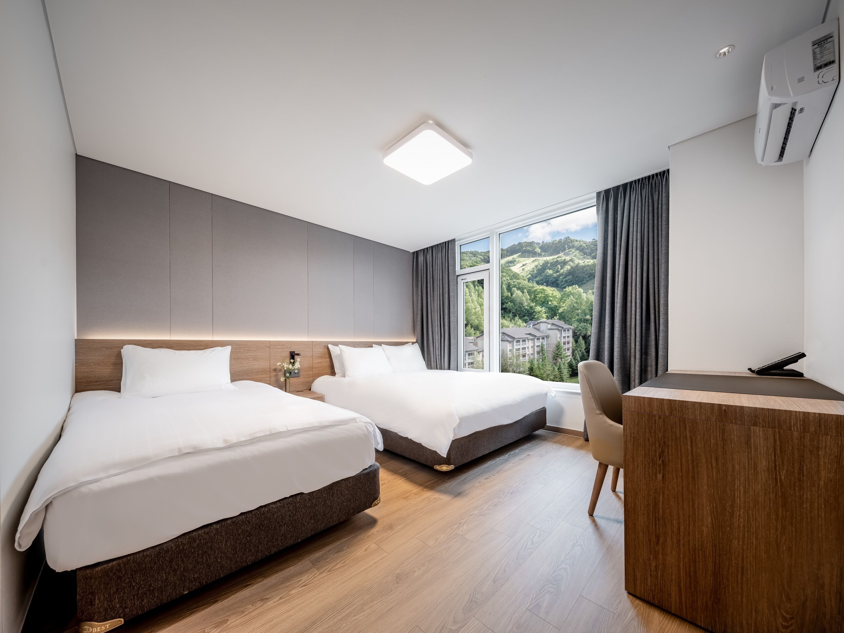 Une chambre d’hôtel moderne, dotée de deux lits, d’un bureau et d’une grande fenêtre donnant sur les collines.