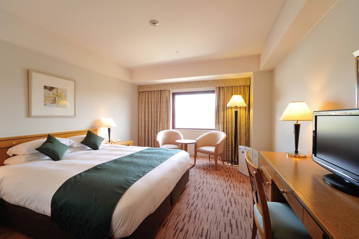 Une chambre d’hôtel avec un grand lit, deux fauteuils, un bureau avec une télévision et une fenêtre avec des rideaux.