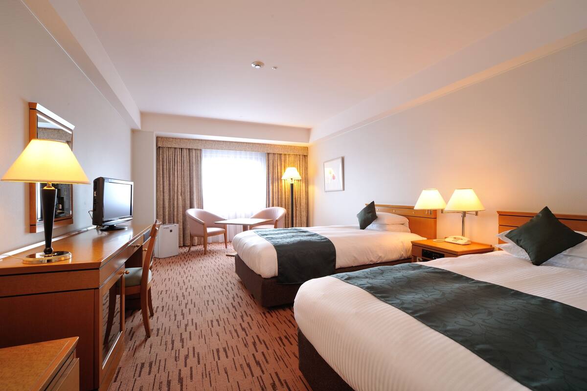 Une chambre d’hôtel avec deux lits, un bureau avec une lampe, une chaise, une télévision et une fenêtre avec des rideaux.