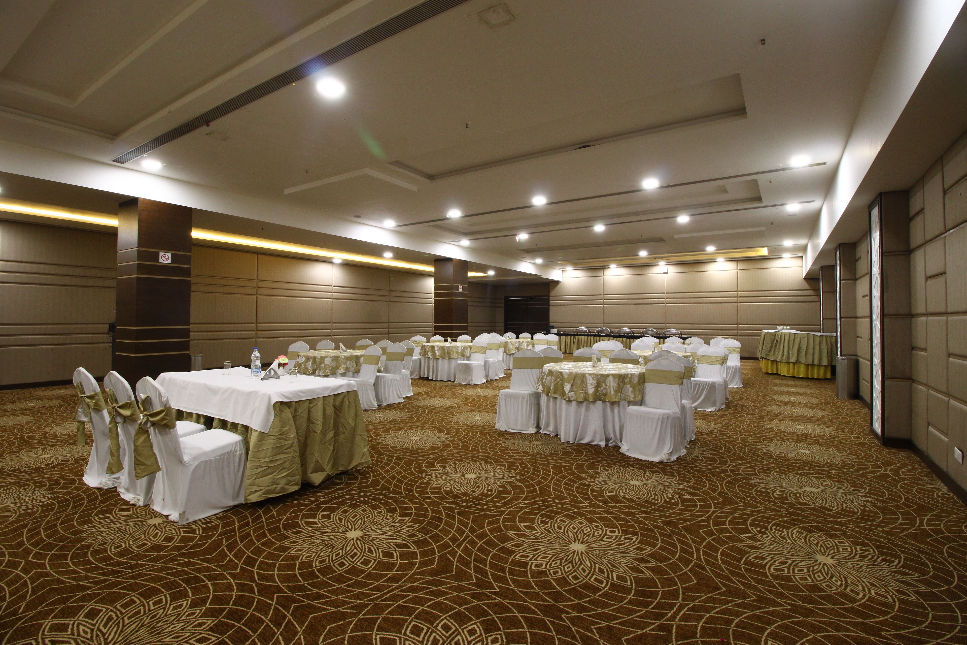Un amplio salón de banquetes bien iluminado con filas de mesas redondas cubiertas con manteles blancos y fajos verdes, dispuesto para un evento formal.